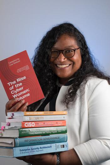 Photo of Michala Liavaag holding a pile of books with the spines showing the titles of each book. She is holding the top book facing the camera; it is a copy of the book 'Rise of the Cyber Women volume 2' - it has a red cover (Michala is a brown woman with long black wavy hair. She is wearing a white jacket.
