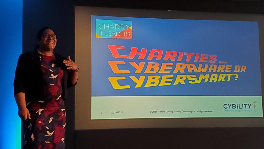 Speaker standing on stage next to a large presentation slide. The smiling presenter, wearing glasses, a dark jacket, and a patterned dress, gestures with one hand while speaking. The slide shows the Charity IT Leaders logo and the bold, colourful title “Charities… Cyber Aware or Cyber Smart?” on a blue background, with the Cybility “Demystifying Cybersecurity” logo along the bottom.