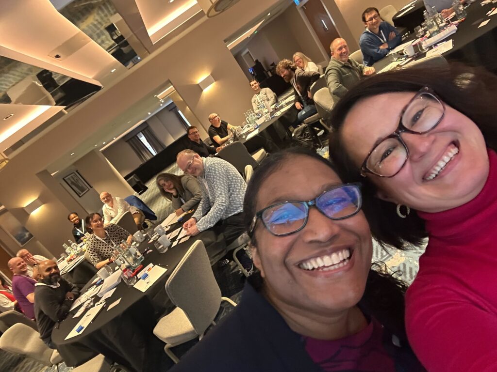 Group selfie taken in a hotel conference room during a workshop. Two smiling facilitators fill the foreground, while participants seated at round tables behind them look towards the camera and smile or wave. The room has mirrored ceiling panels, neutral walls, and scattered papers, bottles, and notebooks on the tables, conveying a friendly, engaged training atmosphere.
