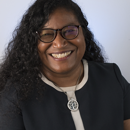 Professional headshot of a person with long curly dark hair and glasses, smiling broadly at the camera against a plain light background. They are wearing a dark dress with a light trim and a round tree‑of‑life pendant necklace.